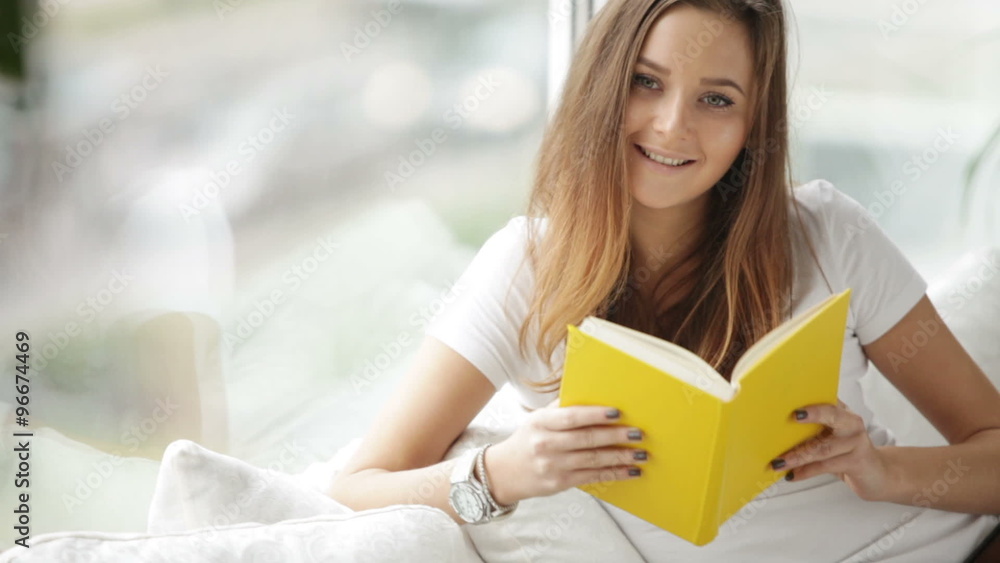 Cute girl sitting by window reading book closing it looking at camera and smiling. Panning camera Cute girl sitting by window reading book closing it looking at camera and smiling. Panning camera