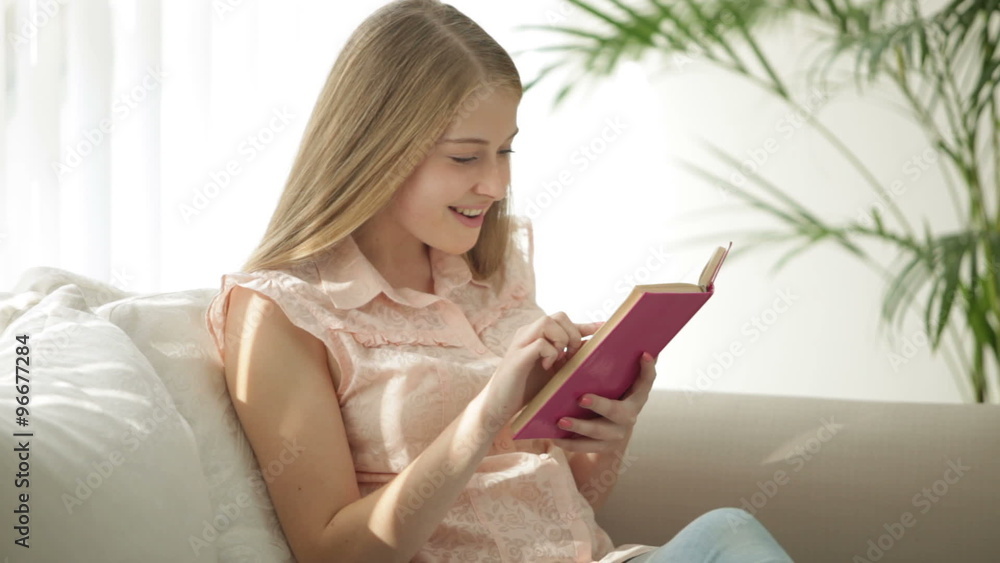 Beautiful girl sitting on sofa reading book looking at camera and smiling. Panning camera Beautiful girl sitting on sofa reading book looking at camera and smiling. Panning camera