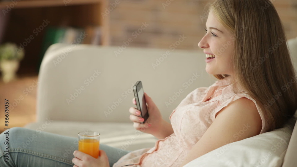 Cute girl sitting on sofa using mobile phone drinking juice and smiling at camera. Panning camera Cute girl sitting on sofa using mobile phone drinking juice and smiling at camera. Panning camera