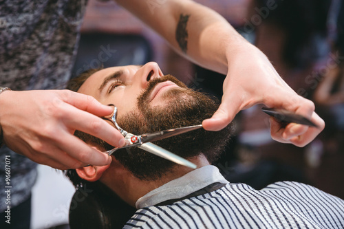 Fotografie Hairdresser doing haircut of beard to young attractive man