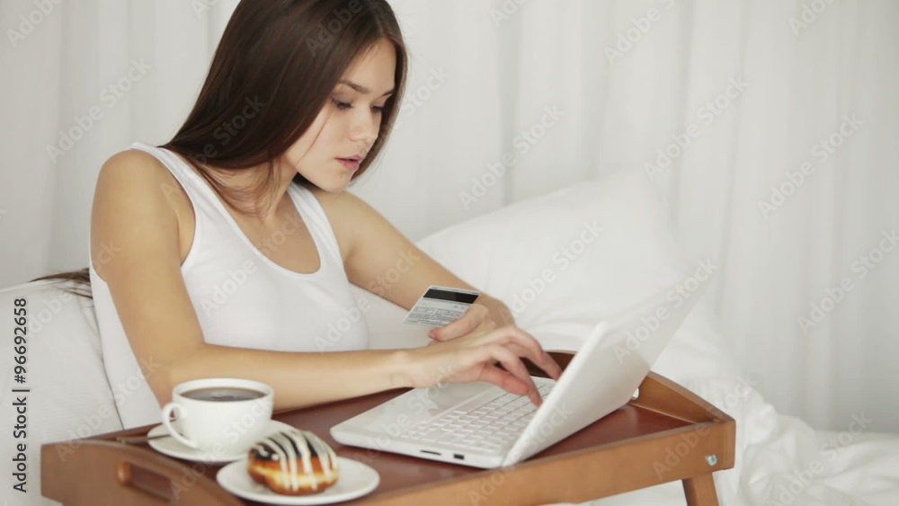Charming girl relaxing in bed using laptop holding credit card looking at camera and smiling. Panning camera Charming girl relaxing in bed using laptop holding credit card looking at camera and smiling. Panning camera