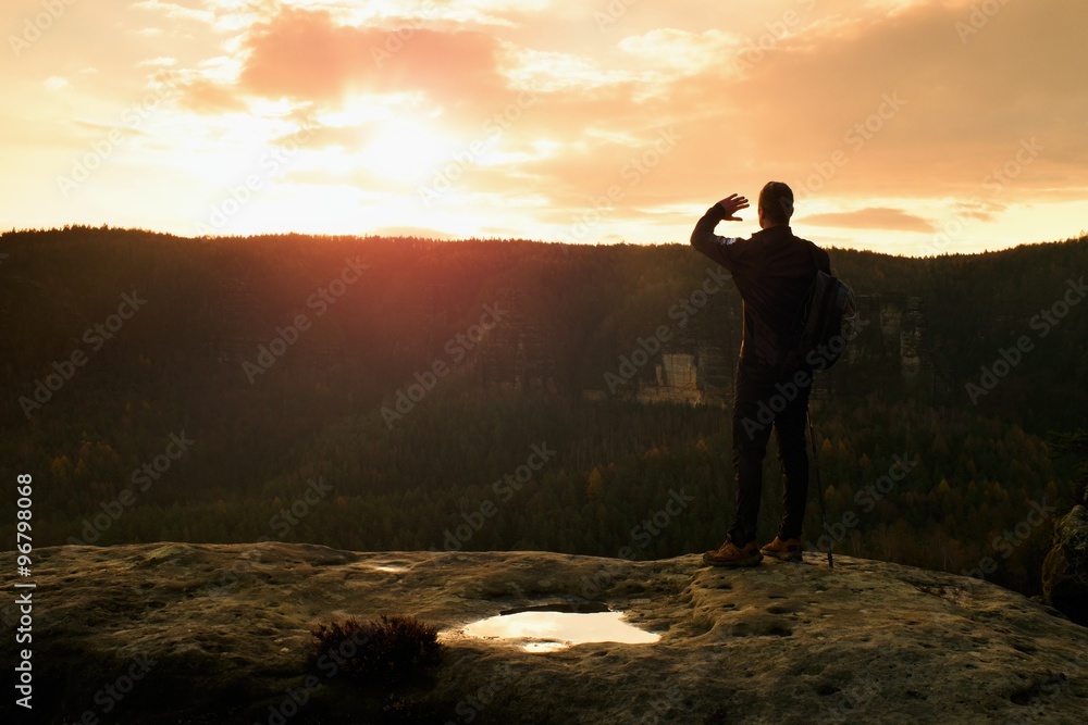 Tourist guide on the way with pole in hand. Hiker with sporty backpack stand on rocky view point above misty valley. Sunny spring daybreak in rocks