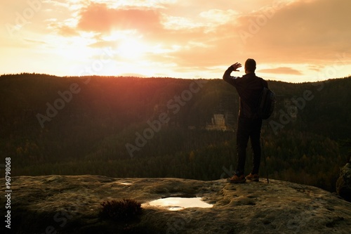 Tourist guide on the way with pole in hand. Hiker with sporty backpack stand on rocky view point above misty valley. Sunny spring daybreak in rocks