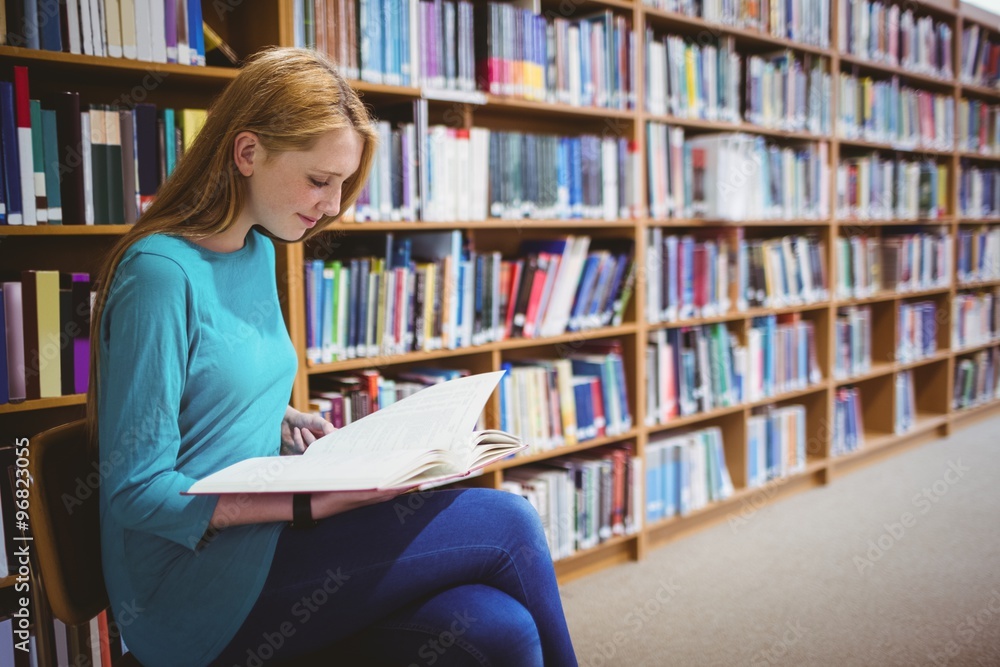 Smiling student sitting on chair reading book in library Smiling student sitting on chair reading book in library