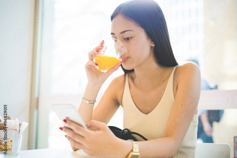Young handsome asiatic long brown straight hair woman sitting in a bar, drinking a juice and using a smartphone, looking downward the screen - technology, social network, relaxing concept Young handsome asiatic long brown straight hair woman sitting in a bar, drinking a juice and using a smartphone, looking downward the screen - technology, social network, relaxing concept