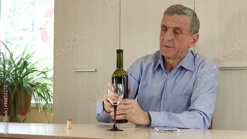 Senior man in blue shirt sits at the table and pours wine in glass. Grey-haired senior man drinks wine. Man with smartphone drinking wine. Man with white mobile phone and bottle of wine
