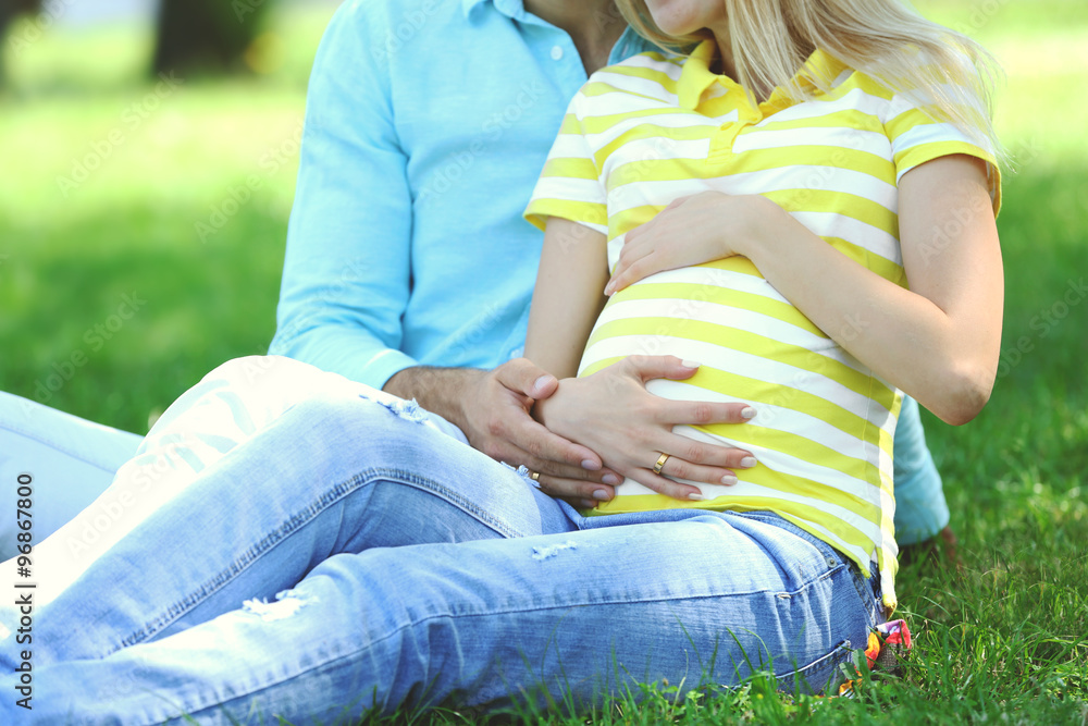 Young pregnant woman with husband sitting on green grass in park