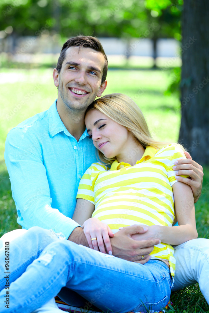 Young pregnant woman with husband sitting on green grass in park Young pregnant woman with husband sitting on green grass in park
