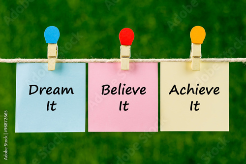 Word quotes of Dream It, Believe It, Achieve It on sticky color papers hanging on rope against blurred green background.