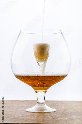 Beer is pouring into a glass which is into another huge glass, creating foam and bubbles on the wooden texture at the bottom and white background