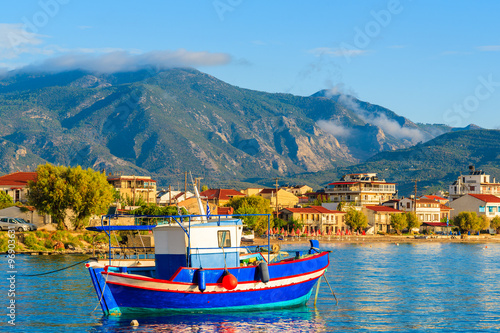 Greek fishing boat on sea with colourful houses in background at sunrise time on Samos island, Greece