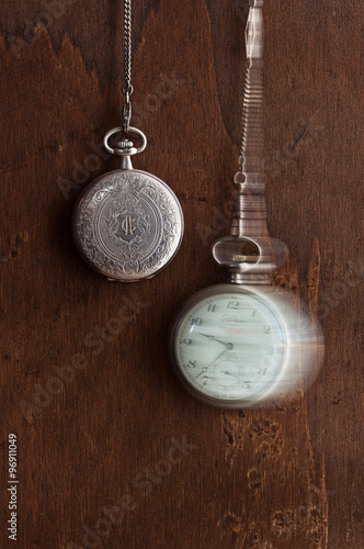due orologi da taschino che pendono dall’alto; uno decorato con incisione e lettere, l’altro oscilla attaccato alla catenella