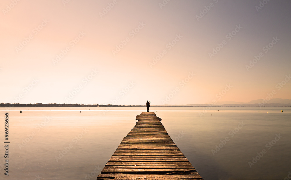 Fototapeta Man standing on a small jetty, enjoying the sunset over a lake