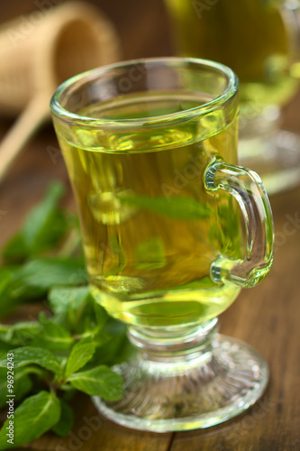 Freshly prepared mint tea out of fresh leaves served in glass cup with leaves on the side and strainer in the back (Selective Focus, Focus on the front rim of the glass)