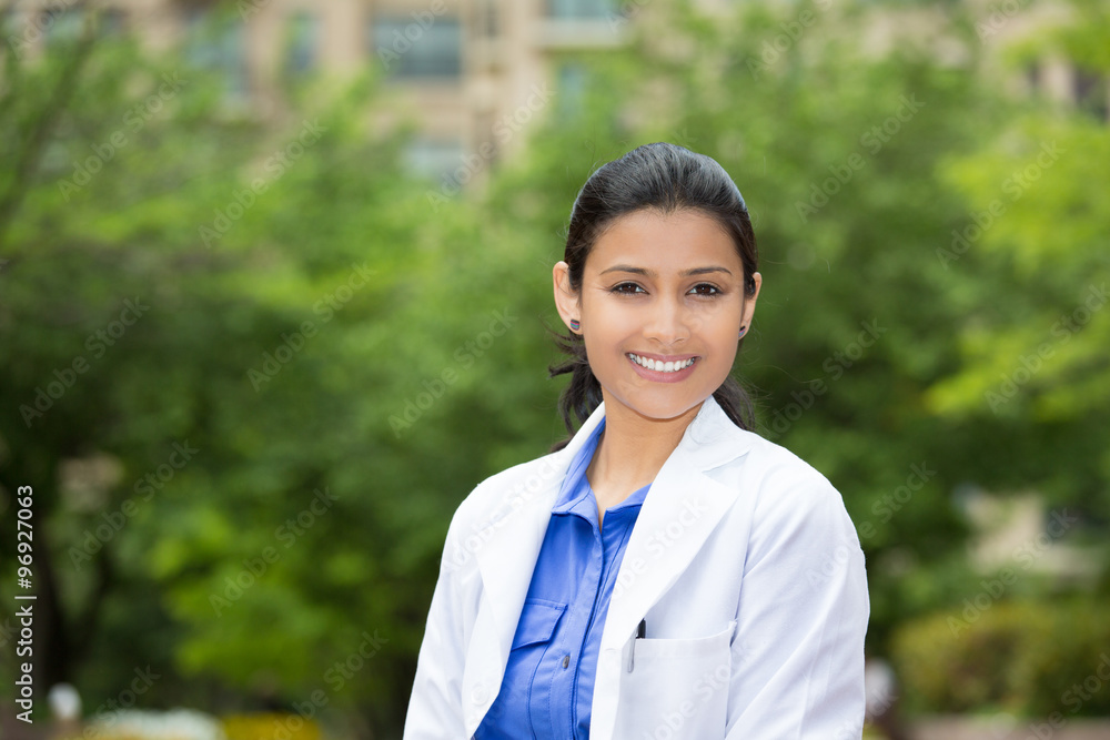 Closeup headshot portrait of friendly, cheerful, smiling confident female, healthcare professional with lab coat. isolated outdoors outside green trees background. Patient visit. Closeup headshot portrait of friendly, cheerful, smiling confident female, healthcare professional with lab coat. isolated outdoors outside green trees background. Patient visit.