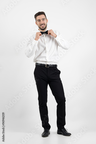 Laughing happy young bearded elegant man adjusting bow tie looking at camera. Full body length portrait over gray studio background.