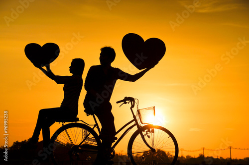 Silhouette of a man and a woman riding a bicycle together and holding big hearts