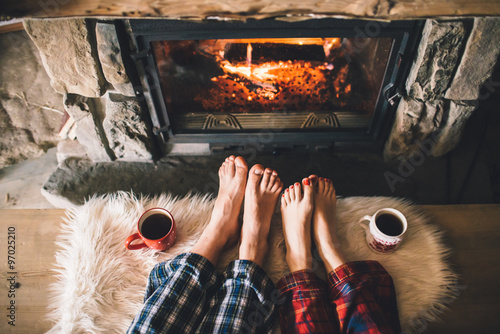 Bare couple feet by the cozy fireplace. Man and Woman relaxes by warm fire with a cup of hot drink and warming up her feet. Close up on feet. Winter and Christmas holidays concept