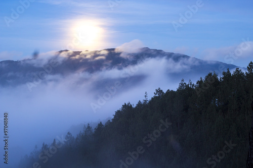 Neblina entre montañas y el bosque acompañados de la Luna