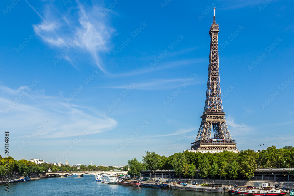 Fototapeta premium Eiffel Tower, Paris, France, September 11, 2015. Shown against a blue sky, with wispy clouds. In the foreground are boats on the river Seine.