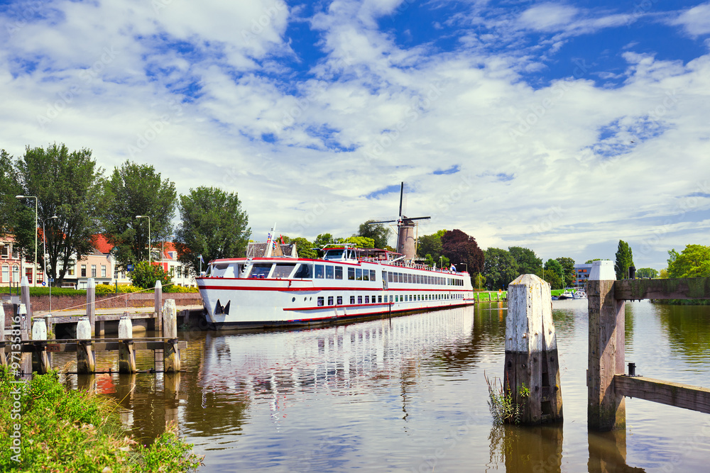 White tour-boat moored in a tranquil canal with a windmill on the background, Gouda, The Netherlands