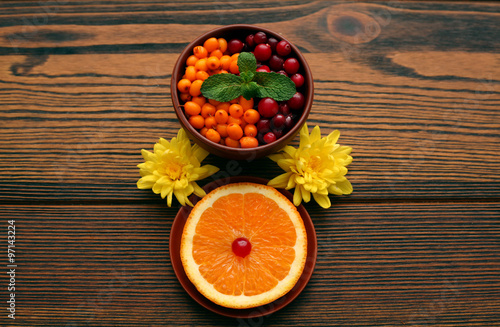 Керамическая чашка с клюквой, облепихой и листком мяты рядом с блюдцем с кусочком апельсина и цветком хризантемы на деревянной доске
