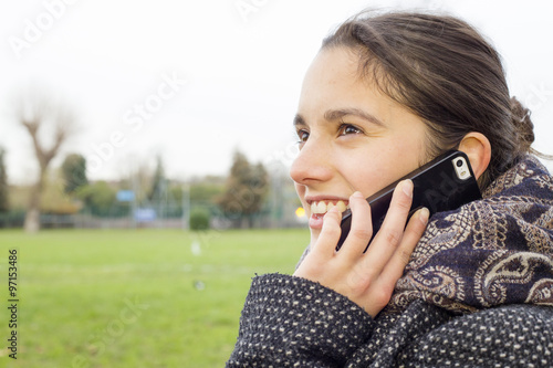 Happy young woman sitting at a bench in a park waiting and answering a phone call. Close up profile shot.