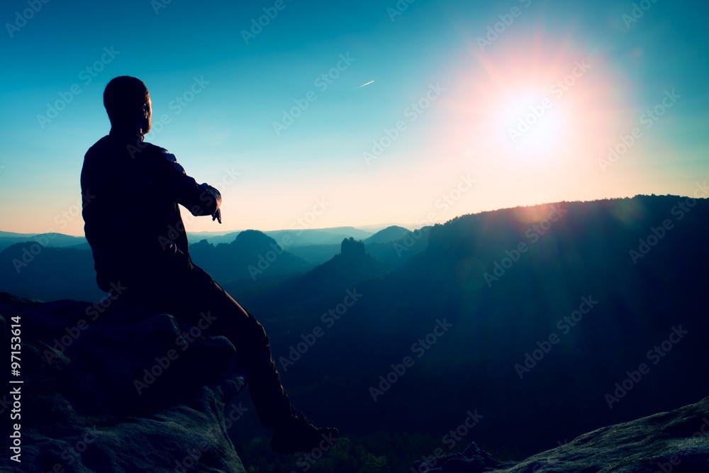 Tourist take a rest. Handsome young man sitting on the rock and enjoying view into misty rocky mountains.