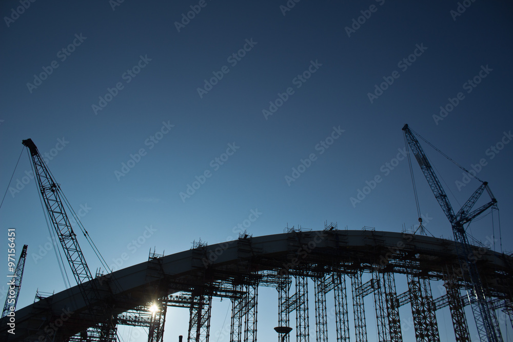Bridge construction - steel arch bridge frame with cranes and girders backlit with sunset light; no people