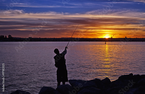 Silhouette of fisherman on the quiet bay with the rays of sunset