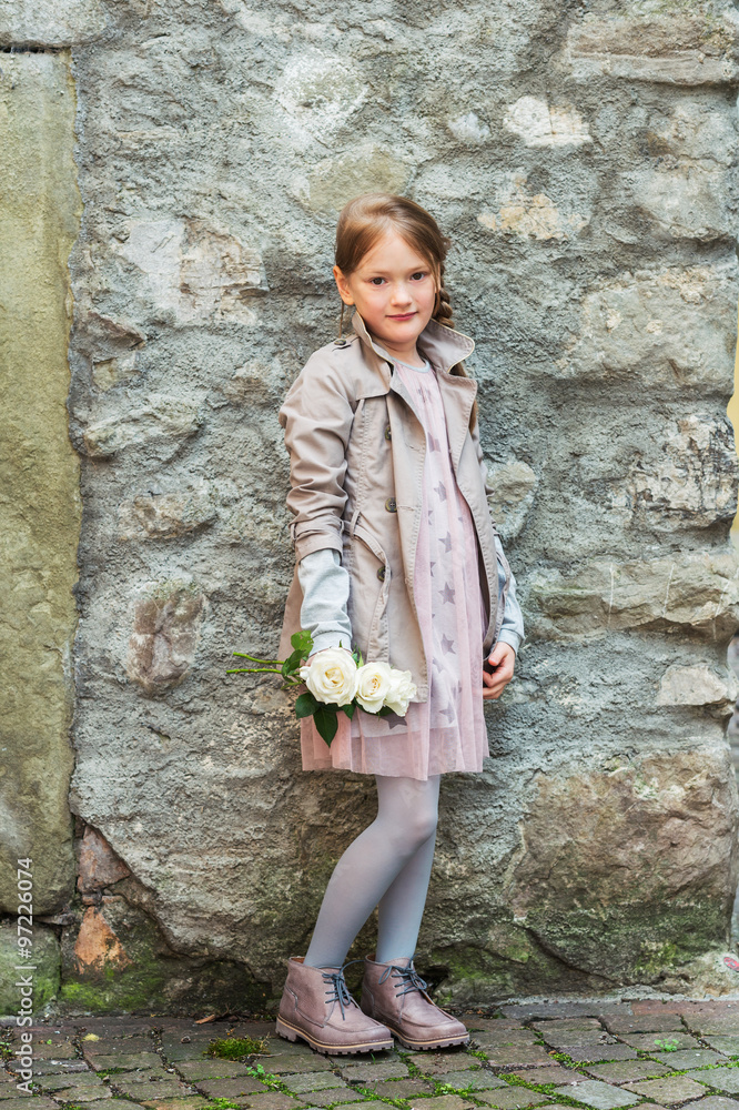 Outdoor portrait of a cute little girl with white roses, wearing beige coat and pink dress Outdoor portrait of a cute little girl with white roses, wearing beige coat and pink dress