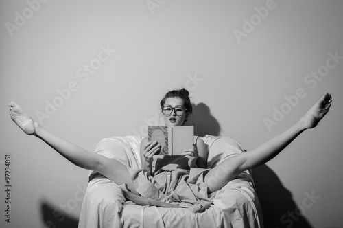Black and white photography of beautiful young lady wearing glasses and holding a book relaxing split legs in chair