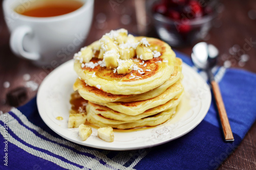 Breakfast: cup of green tea and stack of pancakes with banana slices and coconut flakes on vintage plate on dark blue kitchen towel