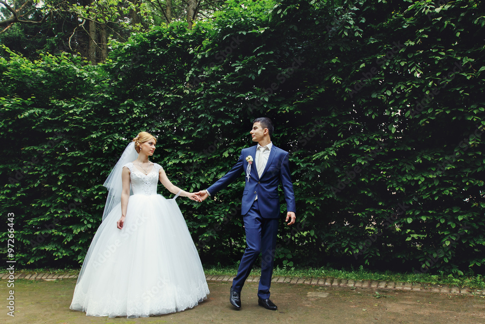 Stylish blonde bride in vintage dress and handsome happy groom holding hands and walking in the park