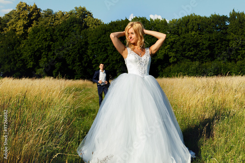Elegant sexual stylish bride posing in the field, with groom and forest in background