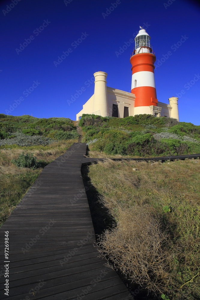 Cape L'Agulhas lighthouse (II) / A lighthouse painted in red and white with a building at the bottom and a wooden boardwalk leading to it. Deep blue and clear sky in the background.
