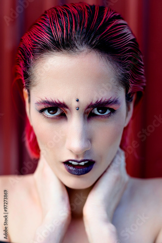 Attractive young woman with colorful makeup  red hairstyle  expressive eyes and piercing on forehead  red metal wall on background