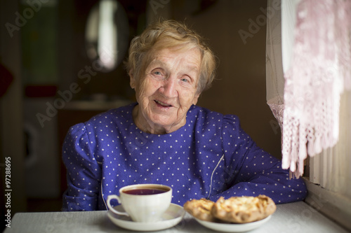 Happy old woman drinking tea sitting at the table in the house.
