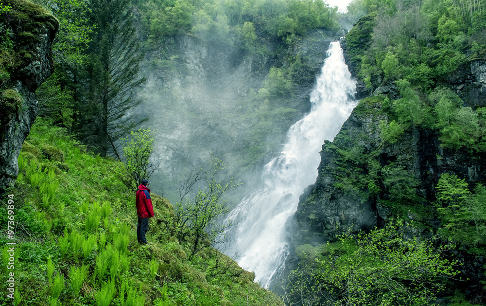 Man standing in front of a large waterfall in the gorge. White water, green trees. Stalheim, Norway. Man standing in front of a large waterfall in the gorge. White water, green trees. Stalheim, Norway.