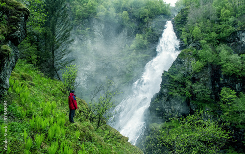 Man standing in front of a large waterfall in the gorge. White water, green trees. Stalheim, Norway.