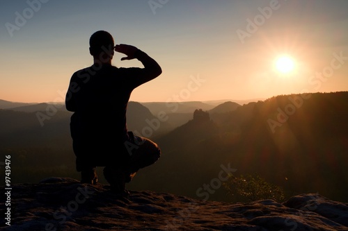 Alone sportsman in black. Tall hiker in squatting position enjoy view at sunset on mountain peak
