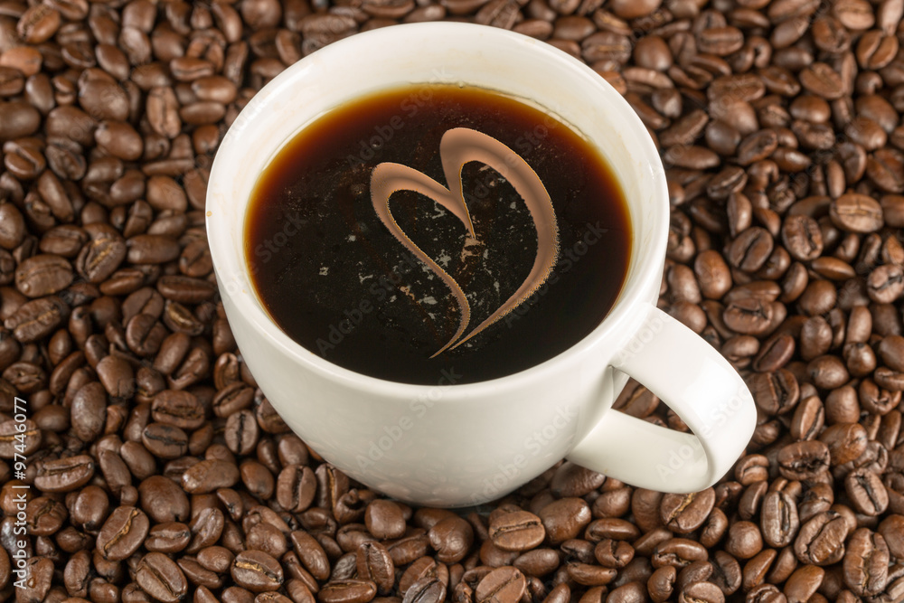 Fototapeta premium Eine weisse Kaffeetasse steht inmitten von Kaffeebohnen. Gefüllt mit Kaffee. Ein geschwungenes Herz verziert den Kaffee.
