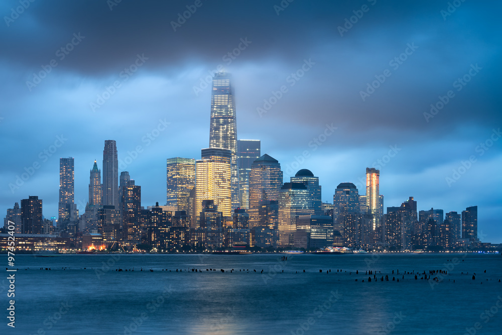 Obraz premium New York City Financial District and illuminated skyline in early evening across Hudson River. Low storm clouds touch the skyscrapers of Lower Manhattan
