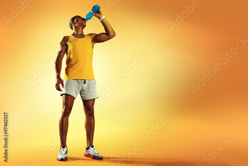 Young muscular build man drinking water of bottle after running, attractive athlete resting after workout outdoors, fitness and healthy lifestyle concept. Isolated on yellow