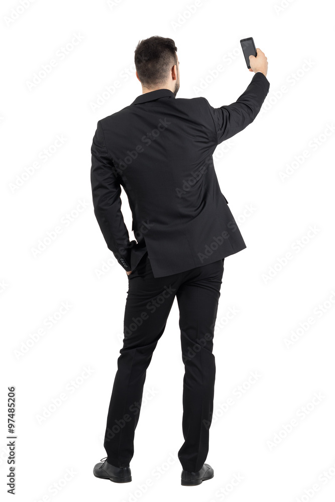 Obraz premium Rear view of young executive in black suit taking photo with his cellphone. Full body length portrait isolated over white studio background. 
