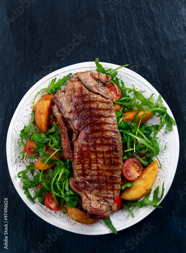 rare beef steak with roasted potatoes, tomatoes and wild rocket leaves. on old blue stone background