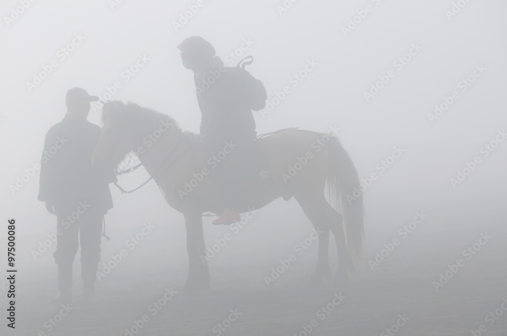 Silhouette of two person and horse in fog and smoke of volcano Bromo