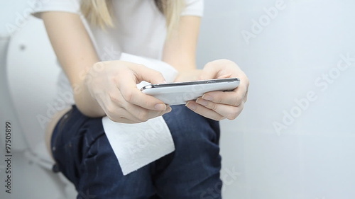 Girl sitting on the toilet using phone in hands .