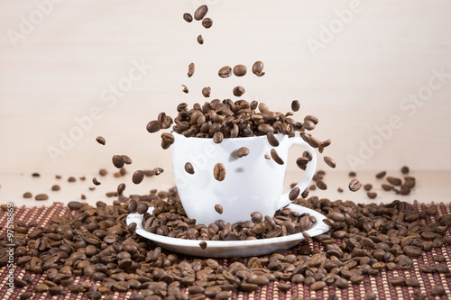 View of white cup standing on white plate with falling down brown roasted coffee beans on tablemat on light wooden textured background.