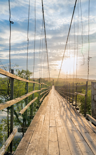 Fotografie, Tablou Old lengthy hanging wooden footbridge with rails over river against sunset background